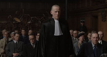 Movie still from “The Last Vermeer” (2019), directed by Dan Friedkin – A man in a black robe stands in front of a group of people; Medium shot, Over the shoulder angle