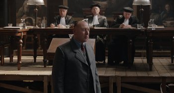 Movie still from “The Last Vermeer” (2019), directed by Dan Friedkin – A man standing in front of a group of judges; Medium shot, Over the shoulder angle