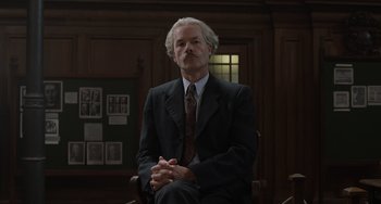 Movie still from “The Last Vermeer” (2019), directed by Dan Friedkin – An older man in a suit and tie sitting in a chair in a room; Medium shot, Low angle
