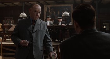 Movie still from “The Last Vermeer” (2019), directed by Dan Friedkin – An older man wearing a suit and tie in front of a group of people; Medium shot, Over the shoulder angle