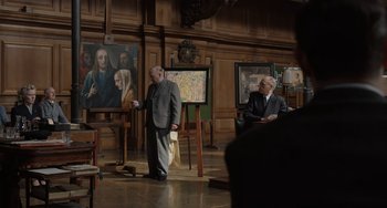 Movie still from “The Last Vermeer” (2019), directed by Dan Friedkin – Two men in suits are standing in front of paintings; Medium shot, Over the shoulder angle