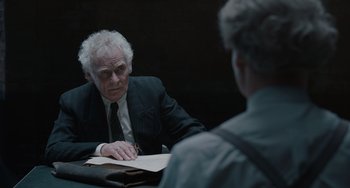 Movie still from “The Last Vermeer” (2019), directed by Dan Friedkin – A man sitting at a table in front of another man holding papers; Medium shot, Over the shoulder angle