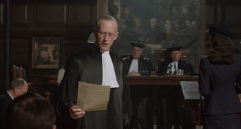 Movie still from “The Last Vermeer” (2019), directed by Dan Friedkin – A man in a black robe holding a piece of paper in a courtroom; Medium shot, Over the shoulder angle