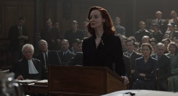 Movie still from “The Last Vermeer” (2019), directed by Dan Friedkin – A woman standing at a podium in front of a group of people; Medium shot, Over the shoulder angle