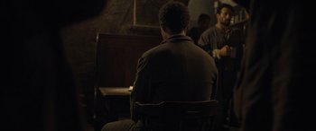 Movie still from “The Last Voyage of the Demeter” (2023), directed by André Øvredal – A man sitting in front of a wooden bench; Medium shot, Over the shoulder angle