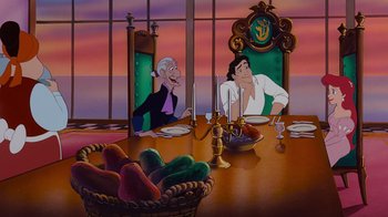 Movie still from “The Little Mermaid” (1989), directed by John Musker – Two people sitting at a dinner table with a basket of fruit on the table; Wide shot, Over the shoulder angle