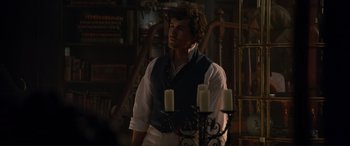 Movie still from “The Little Mermaid” (2023), directed by Rob Marshall – A man standing in front of candles in a dimly - lit room; Medium shot, Over the shoulder angle