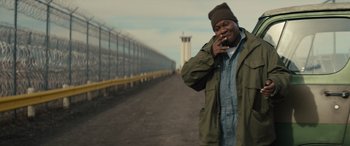 Movie still from “The Locksmith” (2023), directed by Nicolas Harvard – A man smoking a cigarette while standing on the side of a road; Medium shot, Over the shoulder angle