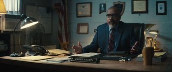 Movie still from “The Locksmith” (2023), directed by Nicolas Harvard – A man sitting at a desk in a suit and tie; Medium shot, Over the shoulder angle