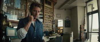 Movie still from “The Locksmith” (2023), directed by Nicolas Harvard – A man talking on a cell phone while standing in a room; Medium shot, Over the shoulder angle