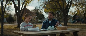 Movie still from “The Locksmith” (2023), directed by Nicolas Harvard – A man sitting at a picnic table with a little girl; Medium shot, Over the shoulder angle