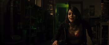 Movie still from “The Locksmith” (2023), directed by Nicolas Harvard – A woman sitting in a dark room smiling for the camera; Medium shot, Over the shoulder angle