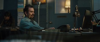 Movie still from “The Locksmith” (2023), directed by Nicolas Harvard – A man sitting at a desk in front of a lamp; Medium shot, Over the shoulder angle