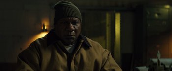 Movie still from “The Locksmith” (2023), directed by Nicolas Harvard – A man wearing a brown jacket and a green hat; Close Up shot, Low angle