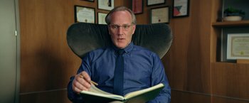 Movie still from “The Machine” (2023), directed by Peter Atencio – A man sitting in an office chair with a notebook; Close Up shot, Low angle