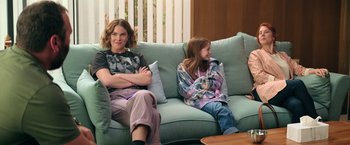 Movie still from “The Machine” (2023), directed by Peter Atencio – A woman and a girl sitting on a couch together; Medium shot, Over the shoulder angle