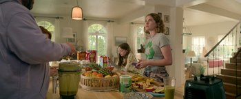 Movie still from “The Machine” (2023), directed by Peter Atencio – A woman and a girl standing in front of a table filled with fruit; Medium shot, Low angle