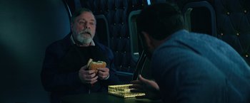 Movie still from “The Machine” (2023), directed by Peter Atencio – A man sitting at a table holding a hot dog in front of another man; Medium shot, Over the shoulder angle