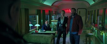 Movie still from “The Machine” (2023), directed by Peter Atencio – A man and a woman standing in a room with a man standing next to them; Medium shot, Low angle