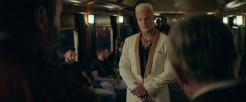 Movie still from “The Machine” (2023), directed by Peter Atencio – A man in a white suit standing on a train; Medium shot, Over the shoulder angle