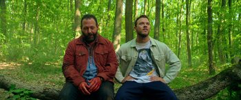Movie still from “The Machine” (2023), directed by Peter Atencio – Two men sitting in the woods , one with blood on his face and the other without; Medium shot, Low angle