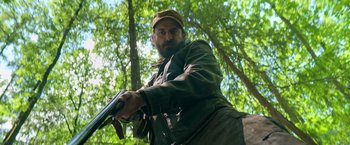 Movie still from “The Machine” (2023), directed by Peter Atencio – A man with a beard holding a gun in the woods; Medium shot, Low angle