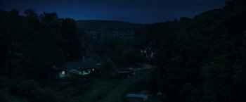 Movie still from “The Machine” (2023), directed by Peter Atencio – A night time view of a forest with a house in the background; Extreme Wide shot, High angle