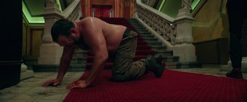 Movie still from “The Machine” (2023), directed by Peter Atencio – A shirtless man kneeling down on the stairs; Medium shot, Low angle
