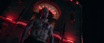 Movie still from “The Machine” (2023), directed by Peter Atencio – A man with tattoos standing in front of a neon sign; Medium shot, Low angle