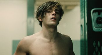 A shirtless man with long brown hair looks upward at the sky