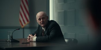 Movie still from “Dopesick” (2021), directed by Danny Strong – A man sitting at a table in front of an american flag; Medium shot, Over the shoulder angle