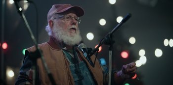 Movie still from “Dopesick” (2021), directed by Danny Strong – An older man with a beard and glasses is speaking into a microphone; Close Up shot, Over the shoulder angle