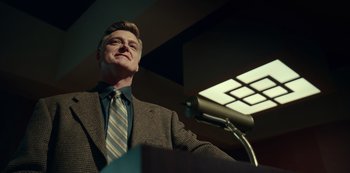 Movie still from “Dopesick” (2021), directed by Danny Strong – A man standing at a podium in front of a microphone; Medium shot, Low angle