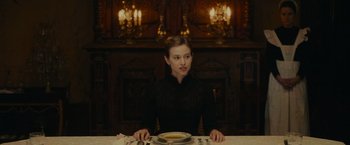 Movie still from “The Mad Women's Ball” (2021), directed by Mélanie Laurent – A woman sitting at a table with a plate in front of her; Medium shot, Over the shoulder angle