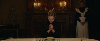 Movie still from “The Mad Women's Ball” (2021), directed by Mélanie Laurent – A woman sitting at a dinner table with her hands clasped in prayer; Medium shot, High angle