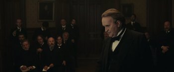 Movie still from “The Mad Women's Ball” (2021), directed by Mélanie Laurent – A man in a suit and bow tie standing in front of a group of people; Medium shot, Over the shoulder angle