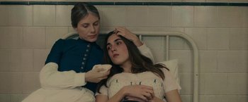 Movie still from “The Mad Women's Ball” (2021), directed by Mélanie Laurent – A young woman is sitting on a bed while a nurse holds a remote control; Close Up shot, High angle