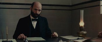 Movie still from “The Mad Women's Ball” (2021), directed by Mélanie Laurent – A man sitting at a table with papers in front of him; Medium shot, Low angle