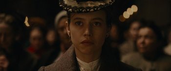 Movie still from “The Mad Women's Ball” (2021), directed by Mélanie Laurent – A young woman wearing a hat in front of a group of people; Close Up shot, Low angle