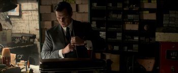 Movie still from “The Man from U.N.C.L.E.” (2015), directed by Guy Ritchie – A man in a suit and tie tying a tie in front of stacks of boxes; Medium shot, Over the shoulder angle