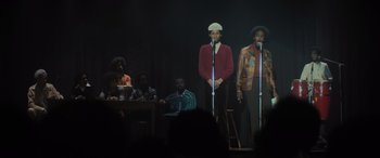 Movie still from “The Many Saints of Newark” (2021), directed by Alan Taylor – A group of people stand on a stage with microphones; Medium shot, Low angle
