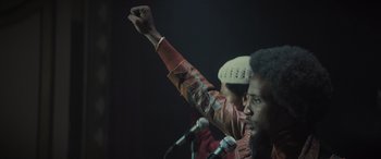 Movie still from “The Many Saints of Newark” (2021), directed by Alan Taylor – A man raises his fist while standing at a microphone; Medium shot, Low angle