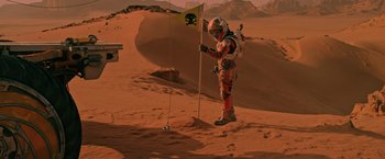 Movie still from “The Martian” (2015), directed by Ridley Scott – A man in a spacesuit standing next to a flag; Wide shot, Low angle