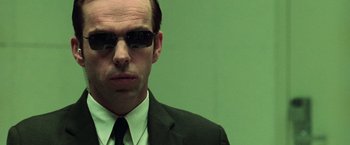 Movie still from “The Matrix” (1999), directed by Lana Wachowski – A man in a suit and tie wearing sunglasses; Close Up shot, Over the shoulder angle