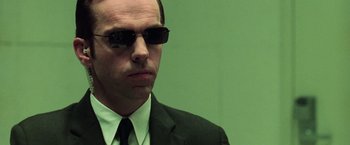 Movie still from “The Matrix” (1999), directed by Lana Wachowski – A man in a suit and tie wearing sunglasses; Close Up shot, Low angle