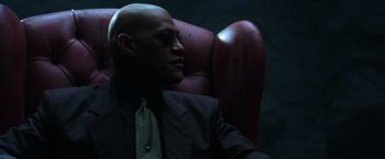 Movie still from “The Matrix” (1999), directed by Lana Wachowski – A man in a suit and tie sitting in a chair; Close Up shot, Low angle