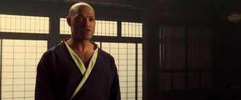 Movie still from “The Matrix” (1999), directed by Lana Wachowski – A bald man in a black and yellow robe stands in front of a window; Close Up shot, Over the shoulder angle