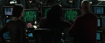Movie still from “The Matrix Reloaded” (2003), directed by Lana Wachowski – A man in a mask sitting in front of two computer monitors; Medium shot, High angle