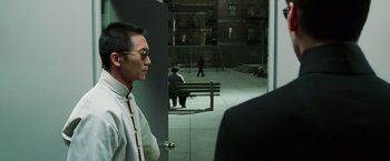 Movie still from “The Matrix Reloaded” (2003), directed by Lana Wachowski – A man wearing a white shirt and a tie standing next to a bench; Medium shot, Over the shoulder angle