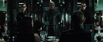 Movie still from “The Matrix Reloaded” (2003), directed by Lana Wachowski – A man standing in front of a plate of food in front of a group of people; Medium shot, Over the shoulder angle